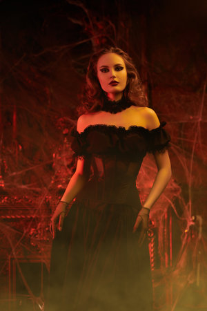 An attractive vampire lady in a low-cut black dress stands in an old abandoned castle in blood red lighting. Sexy witch woman. Halloween. Fairytale vintage style.の写真素材