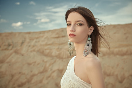 Beauty, Fashion and Boho style. Portrait of a beautiful romantic girl with straight dark hair posing in elegant white dress and boho style jewelry against the sandy valley. Place for text.の写真素材