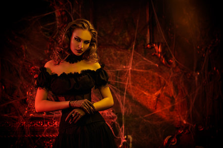 An attractive vampire lady in a low-cut black dress stands in an old abandoned castle in blood red lighting. Sexy witch woman. Halloween. Fairytale vintage style. Place for text.の写真素材