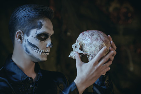 Handsome young man with sugar skull makeup holds a skull in an abandoned castle at Halloween. Dia de los muertos. Day of the dead.の写真素材