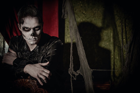Handsome young man with sugar skull makeup poses in an abandoned castle at Halloween. Dia de los...