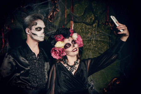 A couple of young people in costumes and make-up of a sugar skull and Calavera Catrina take a selfie in an abandoned castle. A merry Halloween and Day of the Dead celebration.の写真素材