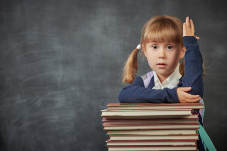 Back to school. Portrait of a little schoolgirl raising her hand in class. Background with black school board and place for text. Education.の写真素材