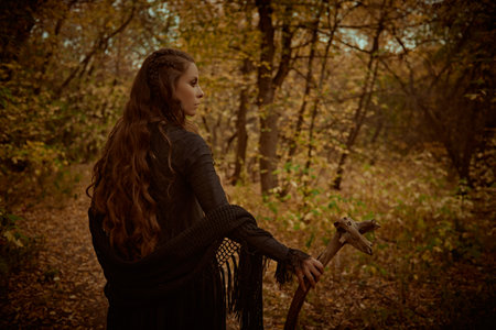 A shaman woman in black garment with ethnic ornaments on her face stands in an autumn forest with a shaman's staff in her hand. Ethnic culture. Shamanic rituals. Copy space.の写真素材