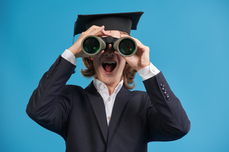 A teenage student boy looks through binoculars. Modern generation. Education. Studio blue background.の写真素材