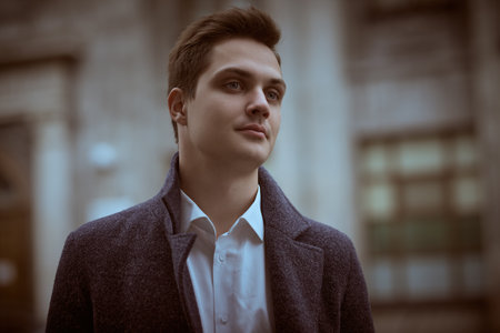 A handsome modern young man in a coat stands on a city street on a cloudy day thoughtfully. People and emotions, lifestyle.の写真素材