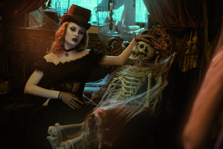 A beautiful and treacherous vampire woman sits in an armchair with a skeleton in an old castle and bares her bloodied fangs. Vampire novels. Vintage style. Halloween.の写真素材