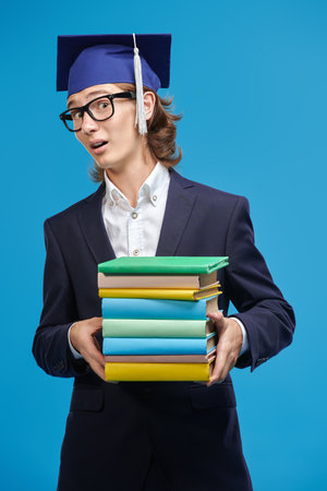 A high school student boy in a suit and academic hat holds books and expresses surprise. High quality education. Successful future. Blue studio background.の写真素材