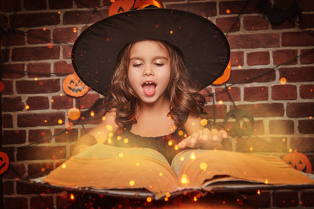 Halloween. Cute little witch casts a spell with a magic wand while standing over a large book of spells.の写真素材