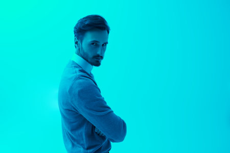 A serious man in a black leather jacket and classic clothes stands in the bright turquoise light and looks seriously to the camera. Feelings, emotions, psychology. Place for text.の写真素材