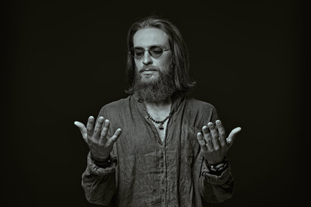 A calm man with a beard and long hair looks at his palms. Black-and-white portrait on a black studio...