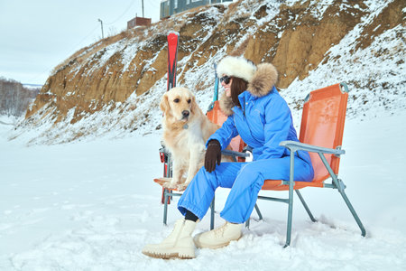 Winter ski resort. A beautiful girl in a blue ski suit has fun playing with her dog, sitting on a mountainside. Snow landscape. Winter holidays.の写真素材