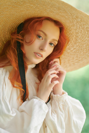A beautiful romantic girl with charming wavy red hair, dressed in a long white shirt and a wide-brimmed straw hat, walks on a sunny summer day. The era of romanticism, 18-19 centuries.の写真素材