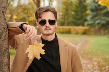 People and lifestyle. A handsome young man in a brown coat stands in an autumn park with a maple leaf in his hand and smiles peacefully. Happy autumn day. Autumn fashion. Copy space.の写真素材