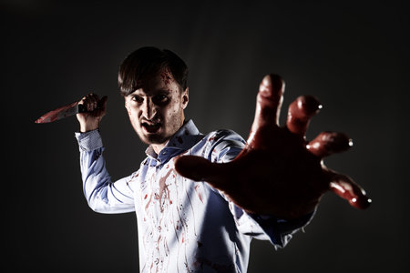 Thriller, horror. A terrible man, a murderous maniac, covered in blood, with a knife in his hand, stretches out his bloody hand to the camera and screams furiously.の写真素材