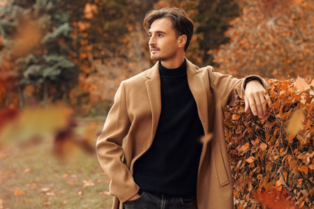 Happy autumn day. A handsome young man in a brown coat stands in an autumn park and smiles peacefully. Autumn fashion. People and lifestyle. Copy space.の写真素材