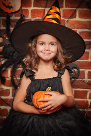 Halloween holiday. Portrait of a little cute girl in a witch costume holding a pumpkin ready for the...