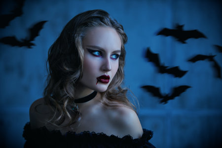 Portrait of a beautiful vampire lady with drops of blood on her lips, standing in a low-cut black dress in a dark room with flying bats. Halloween. Vampires.の写真素材
