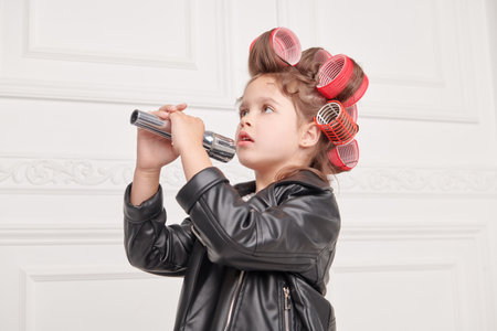 Children's Fashion. Pin-up style. A little fashionista girl in a smart dress with polka dots, in a leather jacket and with curlers on her head is going to a party and singing into a microphone.の写真素材