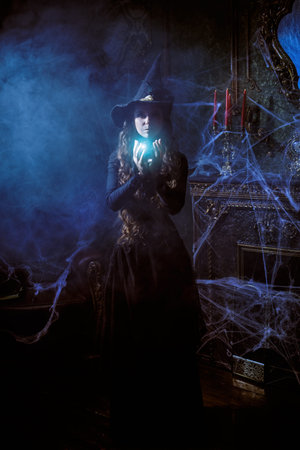 Halloween fairy tales. A beautiful young sorceress in a black hat and dress stands with a shining...