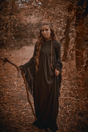 A shaman woman in black garment with ethnic ornaments on her face stands in an autumn forest with a...