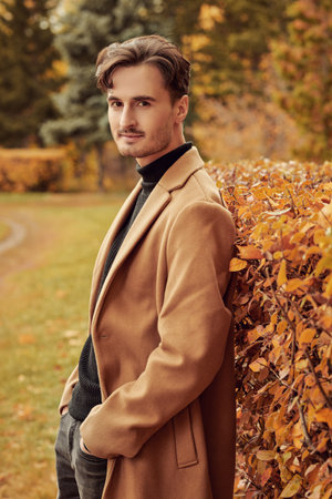 Happy autumn day. A handsome young man in a brown coat stands in an autumn park and smiles peacefully. Autumn fashion. People and lifestyle. Copy space.の写真素材