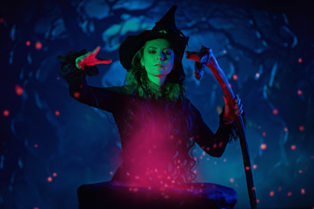 Fantasy, Halloween time. A beautiful young witch in a black dress and hat conjures over a cauldron on a dark moonlit night. Mysterious green and blue smoke is around.の写真素材
