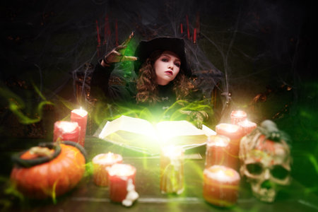 Halloween is a time of magic. A fairytale witch makes magic using a book of spells, sitting in her lair by candlelight. Fantasy.の写真素材