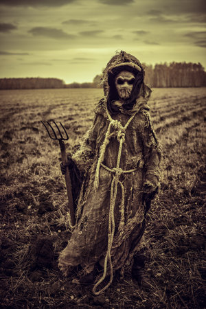 Scary Halloween ghost - a scarecrow with a bag on his head stands alone in a field with a pitchfork in his hand on a gloomy cloudy day.の写真素材