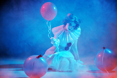 Mysterious old circus. A clown girl in a white circus dress sits on a dark stage, shrouded in haze, and looks intently at the camera, holding a balloon. Halloween.の写真素材