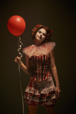 Horror, thriller. Portrait of a wily clown girl dressed in a bloody circus dress, smiling sweetly at the camera and holding a red balloon in her hand. Halloween.の写真素材