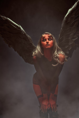 Halloween. Portrait of a seductive devil girl with black wings and bloody horns on a dark studio...