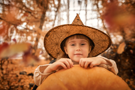 Fairytale children's Halloween. A cute little girl in a witch costume sits by a large pumpkin under the beautiful autumn leaves and is surprised.の写真素材