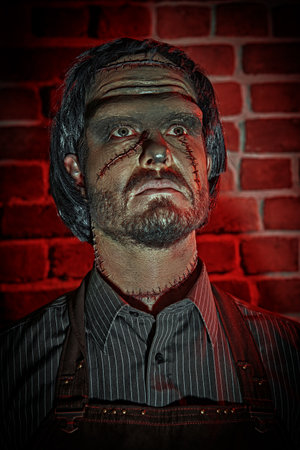 Gothic novel, horror. Halloween. A scary man with scars on his face and neck stands in a dark room with brick walls and a skeleton in the background. Obsessed scientist.の写真素材