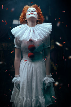 Mysterious old circus. A clown girl in a white circus dress stands on the circus stage, raising her head up to the festive confetti falling on her. Halloween.の写真素材