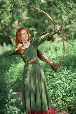 Beautiful young woman archer with magnificent long red hair in a historical celtic dress stands in a forest. Middle Ages. Fantasy. Historical reconstruction.の写真素材