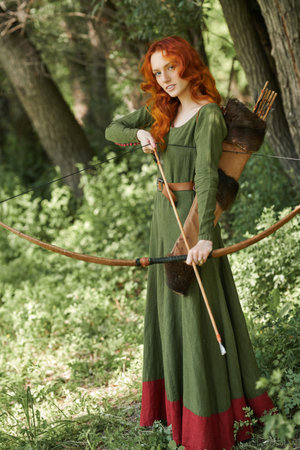 Fantasy. Historical reconstruction. Beautiful young woman archer with magnificent long red hair in a historical celtic dress stands in a forest. Middle Ages.の写真素材