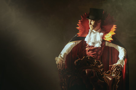 Vampires. An arrogant male vampire in an elegant 19th century style suit stands by a vintage armchair in the dark, illuminated by a beam of light. Halloween. Black background with copy-space.の写真素材