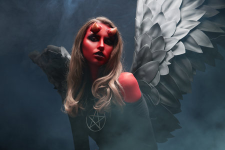 Fantasy heroes. Halloween. An attractive she-devil with bloody horns and black wings, surrounded by haze on a black studio background. Thriller, horror.の写真素材