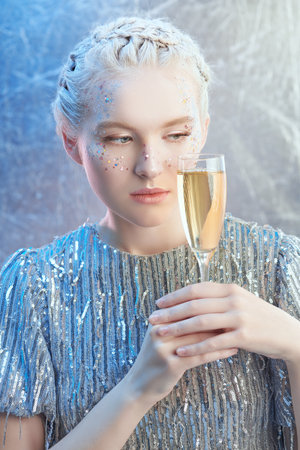Christmas and New Year party look. A beautiful tender girl with light shining make-up and snowy white hair posing in a sparkling dress with a glass of champangne. Winter evening fashion and beauty.の写真素材
