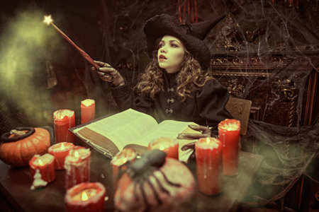Witches' tales. Halloween. A beautiful young witch works magic with a magic wand and a book of spells, sitting in her lair by candlelight.の写真素材