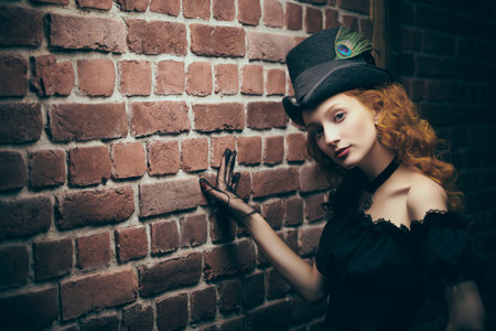 The heroine of the 19th century novel. Beautiful young lady with curly red hair, dressed in elegant black dress with a neckline and a felt hat, standing against a brick wall. Copy space.の写真素材