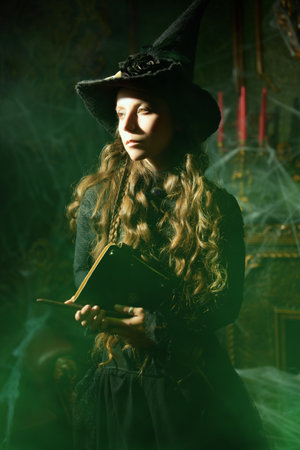 Magic Halloween spells. Beautiful curly-haired sorceress in a black hat makes a spell with a magical book and a wand in a dark old castle covered in cobweb. Art of witchcraft.の写真素材