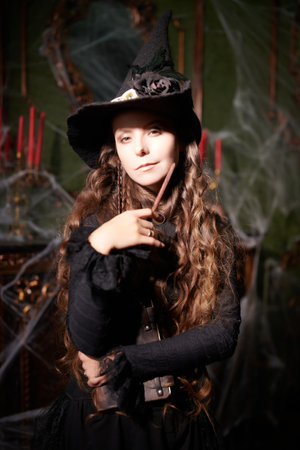 Beautiful curly-haired sorceress in a black hat makes a spell with a magical book and a wand in a dark old castle covered in cobweb. Art of witchcraft. Magic Halloween spells.の写真素材