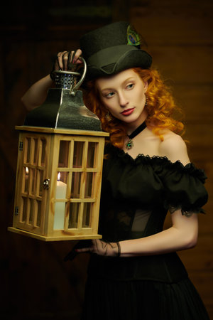 Historical reconstruction of the 19th century. Portrait of a beautiful tender red-haired girl in an elegant black dress with lace and a hat, standing in the light of a lantern. Vintage style.の写真素材