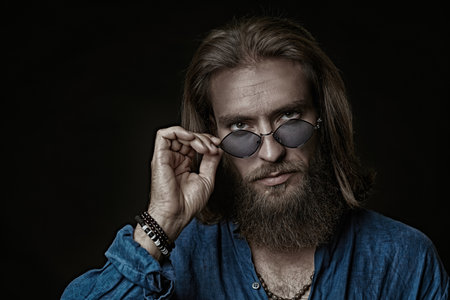 Men's Fashion. A handsome man with a beard and a medium length haircut poses in a natural cotton blue shirt and stylish blue sunglasses on a black studio background.の写真素材