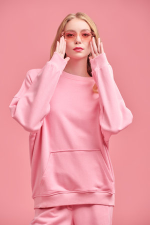 Modern youth feminine style. Beautiful girl fashion model with long wavy blond hair posing in a pink sweatshirt on a pink studio background.の写真素材
