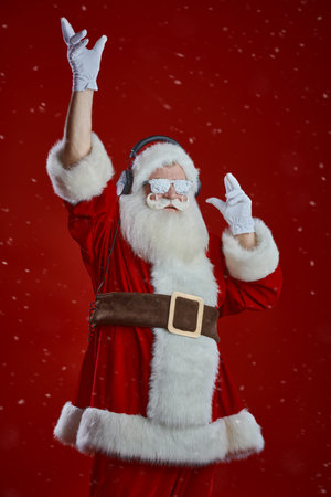 A modern DJ Santa Claus in snow glasses listens to music on headphones and dances. Festive red studio background with snow. Christmas and New Year party.の写真素材