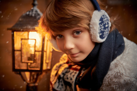 A cute happy boy in winter clothes stands by a lantern at a wooden house and smiles. Fairytale, miracles. Christmas and New Year tales. Winter, snow.の写真素材