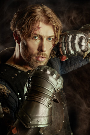 Portrait of a handsome medieval knight with scars on his face in armor, menacingly and boldly looking at the camera. Studio portrait on a black background with a smoke.の写真素材